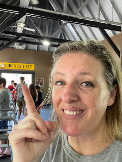 Janet, a white woman with graying blond hair smiles and points to a yellow sign that says