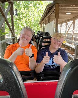 A gray-haired white man in an orange shirt sits next to a strawberry blond white teenager in a blue shirt on a rollercoaster ride. They are both smiling and giving a thumbs up.