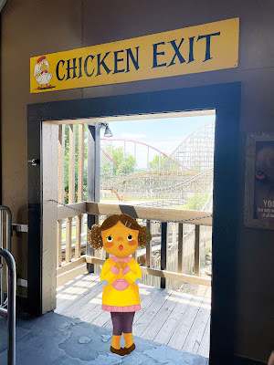 A cartoon of a dark-skinned girl with dark curly hair, wearing a yellow sweater stands in a non-cartoon photograph of a doorway with a rollercoaster in the background. Above the doorway is a sign that says