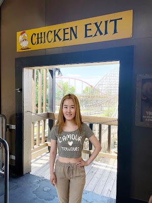 Eunji, an Asian girl with bleached hair wearing brown slacks and a gray shirt, stands in a doorway with a rollercoaster in the background. Above the doorway is a sign that says