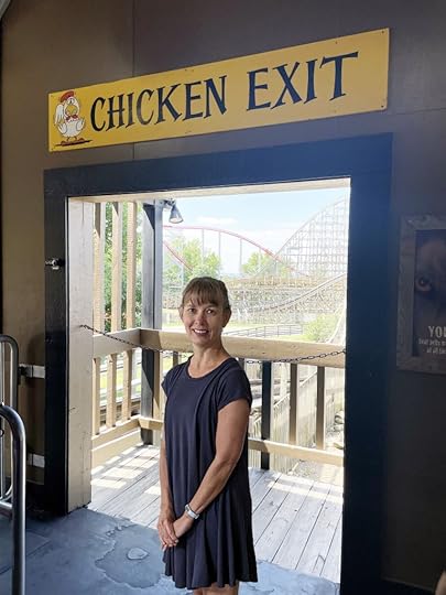 Trish, a white woman with light brown hair wearing a blue dress, stands in an open doorway with a rollercoaster in the background. Above the door is a yellow sign that says 