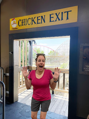 A white woman with dark hair in a pink skirt and navy shorts stands in a doorway with a rollercoaster in the background. Above the doorway is a sign that says