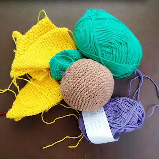 A group of small crocheted items in yellow, brown and green and two balls of cotton yarn - one green and one purple. It isn't clear what the objects are.