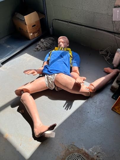 A manniquin in a blue t-shirt and white shorts, with a yellow climbing strap around his torso, is lying on a grey concrete floor, with limbs twisted sll different ways, as though just thrown down. It is lying in a shaft of sunlight. Image taken by Patrick Mcdonaugh