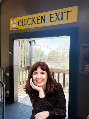 A white woman with dark hair in a dark, flower-print shirt sits in a doorway with a rollercoaster in the background. Above the doorway is a sign that says