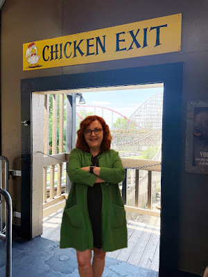 A white woman with red hair in a green coat and black dress stands in a doorway with a rollercoaster in the background. Above the doorway is a sign that says
