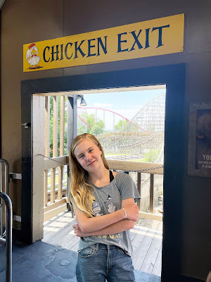 A white teenaged girl with blond hair, wearing jeans and a t-shirt stands in a doorway with a rollercoaster in the background. Above the doorway is a sign that says
