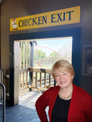 Janet, a light-skinned woman with light brown hair in a red shirt stands in a doorway with a rollercoaster in the background. Above the door-frame is a yellow sign that reads