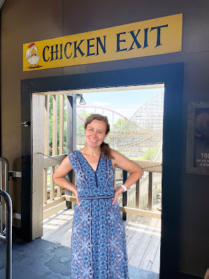 A white woman with dark hair in a blue patterened dress stands in a doorway with a rollercoaster in the background. Above the doorway is a sign that says
