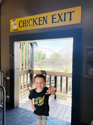A white boy with brown hair in a black shirt and camo pants stands in a doorway with a rollercoaster in the background. Above the doorway is a sign that says