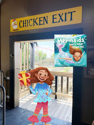 A cartoon girl with red hair and light brown skin, wearing a blue shirt and red striped pants and holding a wrapped present, stands in front of a real life door with a rollercoaster in the background. Above the door is a yellow sign that reads