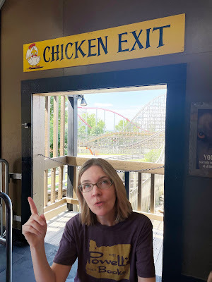 A light-skinned woman with blond hair in a dark grey shirt stands in a doorway with a rollercoaster in the background. Above the doorway is a sign that says