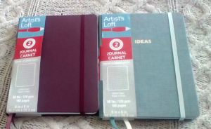 Two Artist's Loft series two notebooks are side by side on an Irish fisherman knit afghan. One notebook is a wine-colored faux leather cover, and the other is sage green cloth, with 