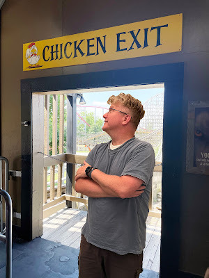 A light-skinned man with gray-blond hair wearing a light blue t-shirt and dark shorts is standing in front of a doorway with a rollercoaster in the background. Above the doorway is a yellow sign that says