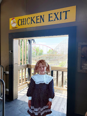 A light-skinned, blond girl in a blue dress with white lace, stands in front of a doorway with a rollercoaster in the background. Above the door is a sign that says