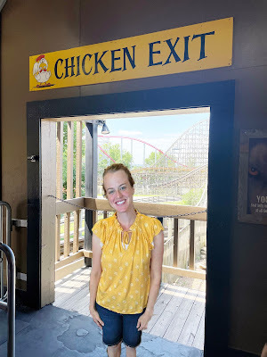a light-skinned woman with blond hair in a yellow shirt and blue shorts is standing in front of an open doorway with a rollercoaster in the background. Over the doorway is a yellow sign that says