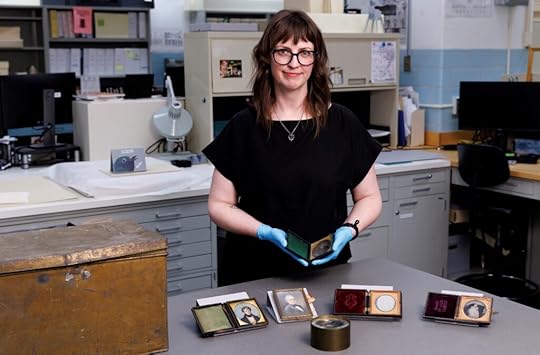 Color photo of woman with glasses, dressed in black, wearing blue preservation gloves, standing in a lab. On a table in front of her are several framed photos. She holds one in her hands.