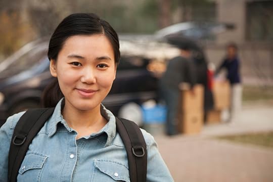 Young woman is smiling and wearing chambray shirt with black backpack. In background her family is unloading dorm room essentials from minivan.
