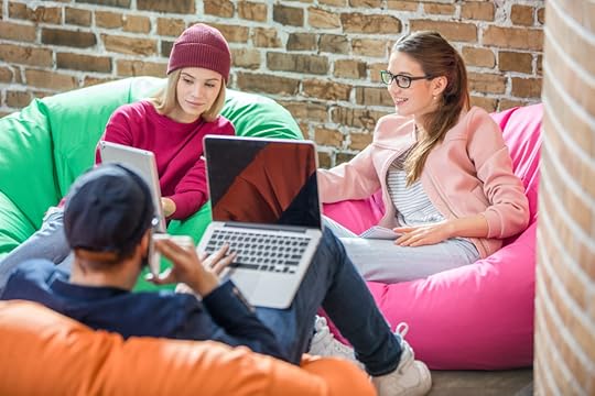 A young man sits in orange bean bag with his laptop computer open. Two young women face him. Both are sitting in bean bags and sharing an electronic tablet.