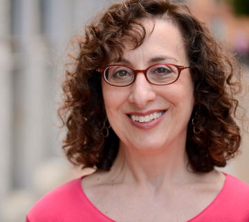 Headshot of Deborah Sosin smiling. She is wearing a pink top, with red glasses on and looking at the camera.