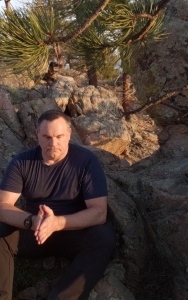 The author meditates in a peaceful mountain setting shortly before sunset. A man sitting with his hands together on a rock outcrop under a pine tree (Photo by TJ Burr, 5/7/2021).