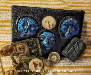 Elder Futhark rune Ansuz on wrought iron, with Isa and Thurisaz in beige polymer clay with dried rosemary, on a mixed media repreentation of the Norns with bright blue faces on a dark small canvas, a slice of reindeer antler, and small trumpet mushrooms preserved in acrylic, on a striped copper and cream tea towel.