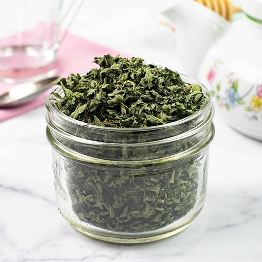 A small glass jar filled to overflowing with dried mint, a tea pot, spoon and napkin in background.