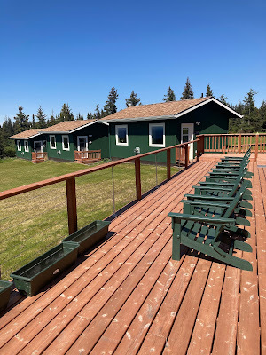 View of a back patio to main cabin, with six green adirondack chairs and empty planter boxes. Two green cabins with white trim and brown roofs in mid-background. Landscape of pines in distant background.