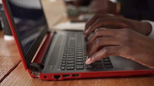 A pair of brown-skinned hands type on a black and red laptop sitting on a wood desk.