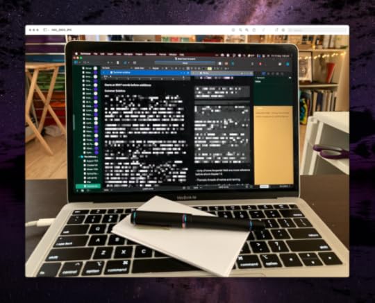 A laptop with a black fountain pen with shimmery metal lying on the keyboard on top of some index cards. The screen shows a Scrivener window with multiple areas of text and notes, in shades of dark green, black, and gold backgrounds. The content of the screen is described in the following text.