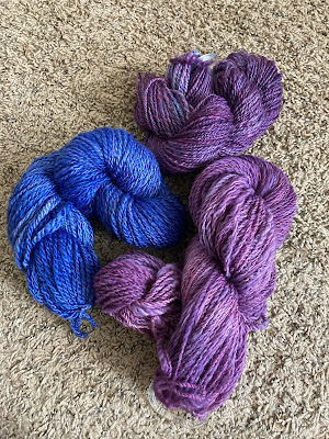 three hanks of homespun, hand-dyed yarn: one a deep, vibrant blue-purple, one a medium purple with tonal variations, and one a darker purple tonal variations leaking into the blue range.