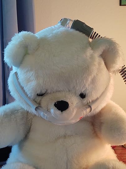 Stuff white toy bear is shown wearing a CPAP mask with straps around his head and a hose coming out the top of his head to demonstrate how it looks.