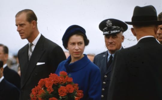 Queen Elizabeth, in a blue dress and hat, holding roses, with Prince Philip to her right and two U.S. military officers on her left, apparently just after arrival.