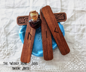 Elder Futhark runes Kenaz, Isa, and Urus carved into cedar, with a small heart-shaped vial of honey, resting above a blue dish of water, all on an offwhite linen background