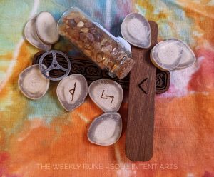 Elder Futhark runes Kenaz carved into cedar, Jera, and Thurisaz reversed carved into reindeer antler slices, with a small vial of baltic amber and a metal wheel with antler slices scattered on a rainbow ice-dyed background