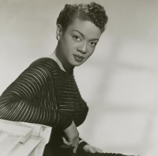 Hazel Scott in a black and white publicity photo, wearing a black dress, seated at the edge of a white piano