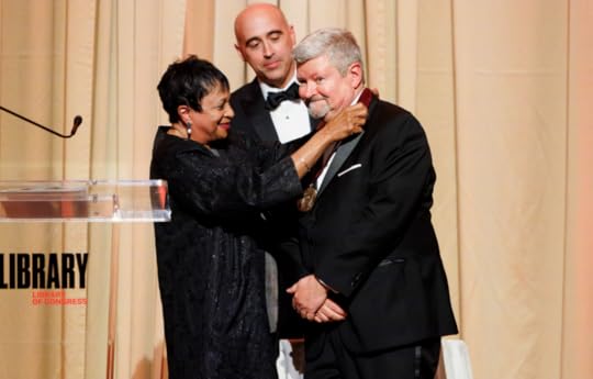Color medium shot of Librarian of Congress Carla Hayden bestowing the Kluge Medal on George Chauncey, while Kluge Director Kevin Butterfield looks on. Both men are wearing black tuxedos; the Librarian is wearing a black dress. The on-stage curtains behind them are a light gold.