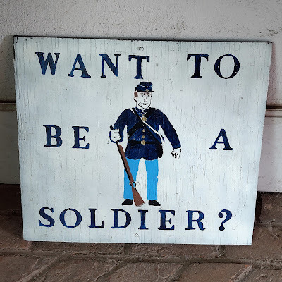 A wooden sign with a soldier drawn on it that says, "Want to be a soldier?"