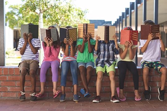 A variety of students sits on a low brick wall at a school, reading open books they each hold up in front of their faces. Many kids are playing catch-up with education and covid-19.