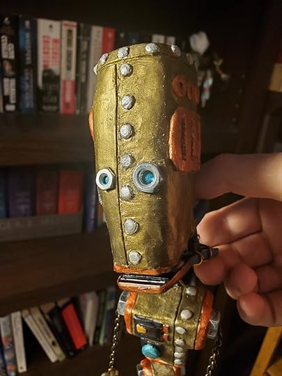 Polymer clay robot puppet painted with shiny metalic paints of bronze, copper, and silver. Robot is facing camera, mouth open, with a hand holding it from behind. By Andy Reynolds.