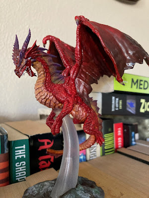 a dragon miniature, painted red, and balanced on top of some books on a shelf.