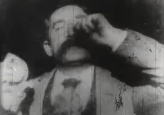 A grainy black and white image of a head-and-shoulders shot of a man wearing a suit in the early stages of a sneeze
