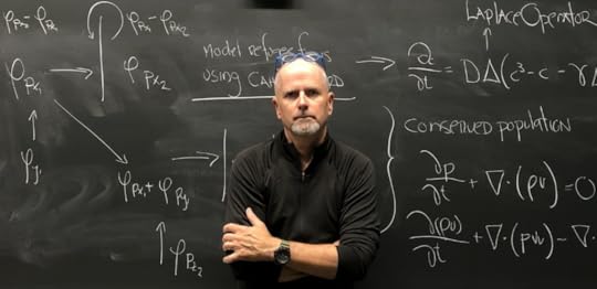 John Hessler, arms folded, stands in front of a blackboard filled with math calcuations written in white chalk