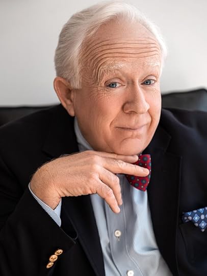 Portrait color photo Leslie Jordan in a black suitcoat with a blue shirt, red bow tie and bright blue pocket square. He is smiling with his lips closed, eyebrows raised, right hand and extended fingers under his chin.