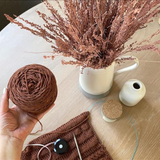 textured knit in browns laying on table with tape measure and Shannon's hand holding a brown skein of wool, with a vase and pitcher of dried flowers on table.