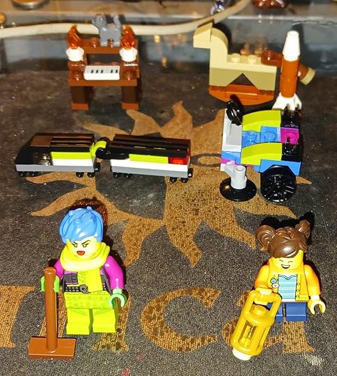 Front row: Two Lego minifigures; back row: four Lego mini-models, an organ with a cat on top, a rocking horse with a rocket, a train engine and car, and a Zamboni