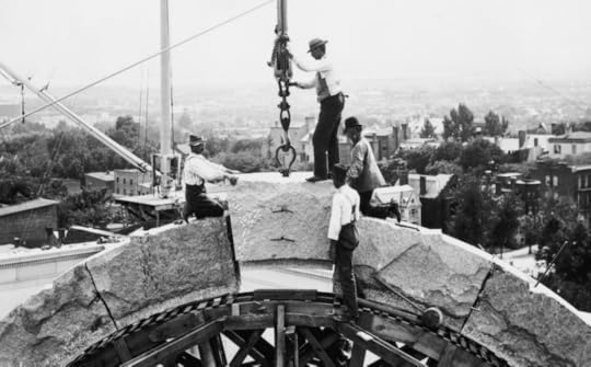 Three workers atop an arch, high on the Library building, as a cran lowers a massive stone block into place. The Capitol Hill neighborhood is visible in the background.