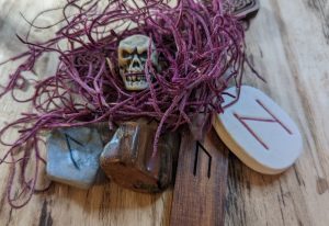 Elder Futhark runes Hagalaz carved into a white ceramic tile, Uruz carved into a cedar stave, and Laguz carved into silver polymer clay tile, with a brownish red chunk of petrofied wood, a bit of dried red moss above, and a small plastic skull on a light wood-grained background