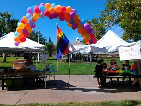 Photo of a Pride festival with a balloon banner and a pride flag in the center and booth canopies on either side.