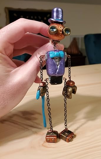 A metalic-painted robot puppet made by Andy Reynolds, made of clay and chain, with a clip for the mouth. It has a copper head and bronze eye scopes and mouth. It's wearing a purple tophat, a purple suit, a teal bowtie and a teal cane.